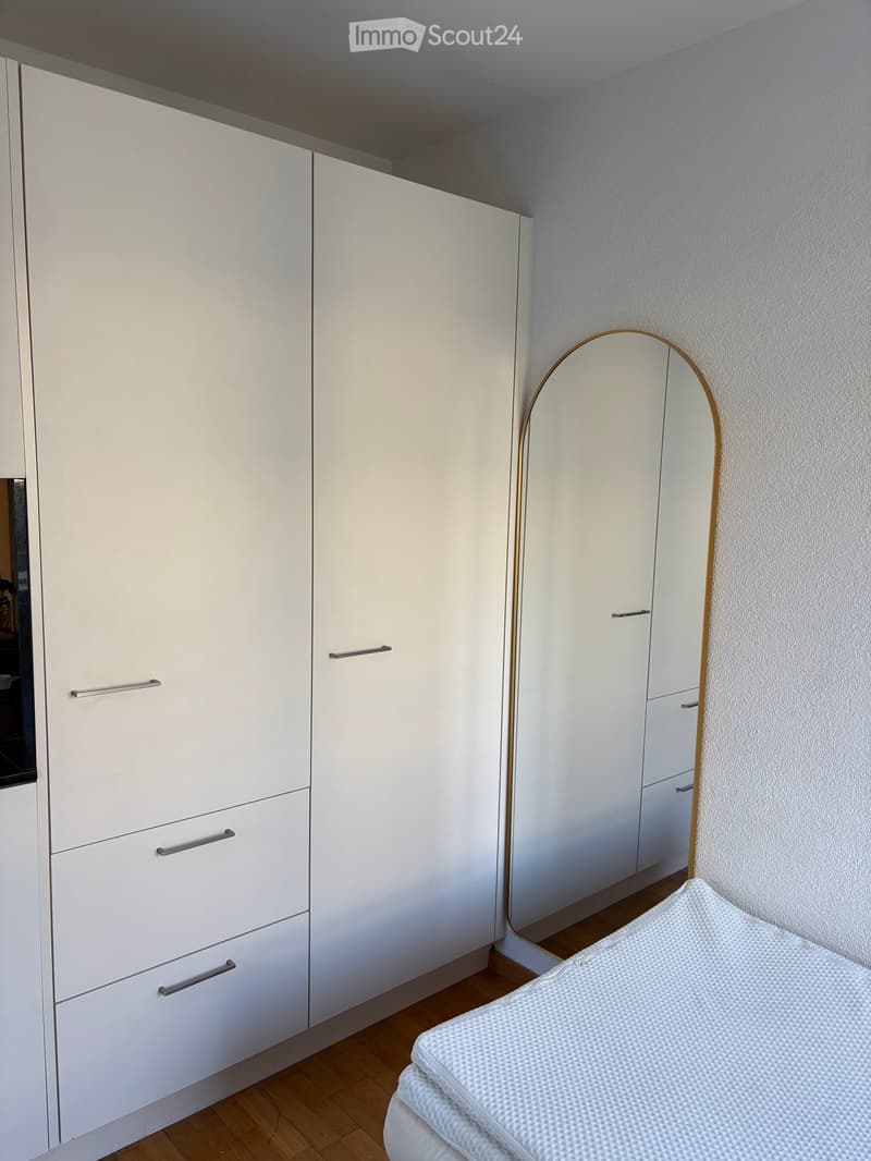 Modern bedroom with white built-in wardrobe, a round mirror with a gold frame, and a neatly made bed with white bedding.