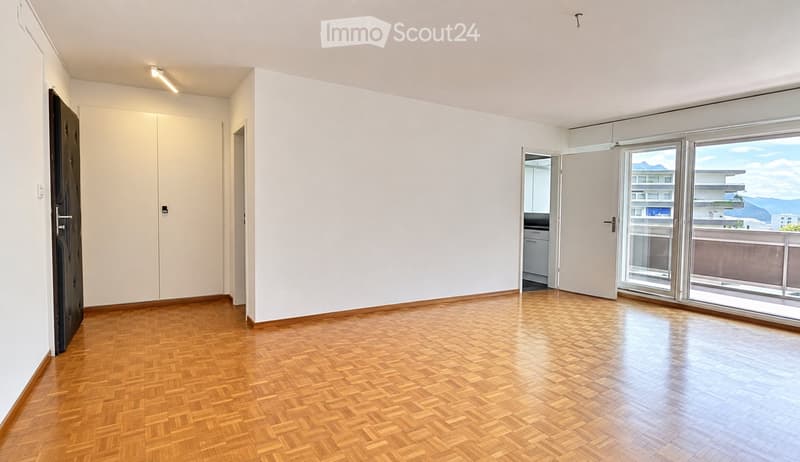Empty room with wooden herringbone flooring, white walls, large windows, and a door leading to a balcony.