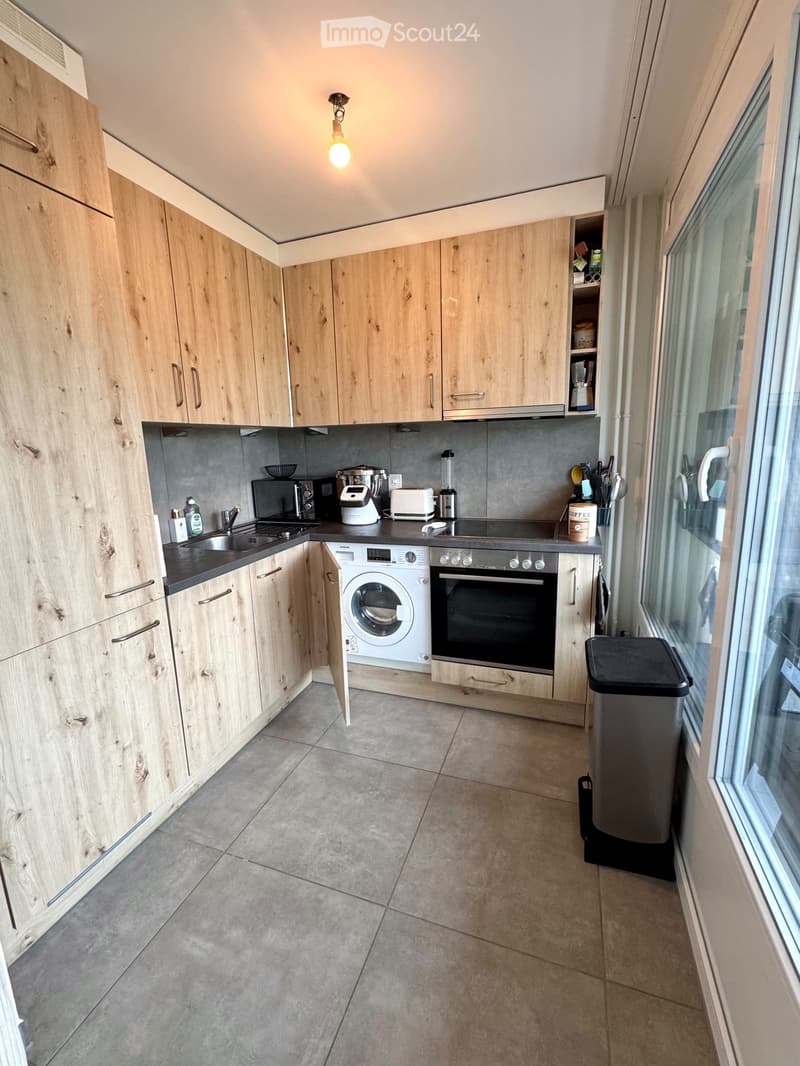 Modern kitchen with light wooden cabinets, stainless steel appliances, and a tiled floor.