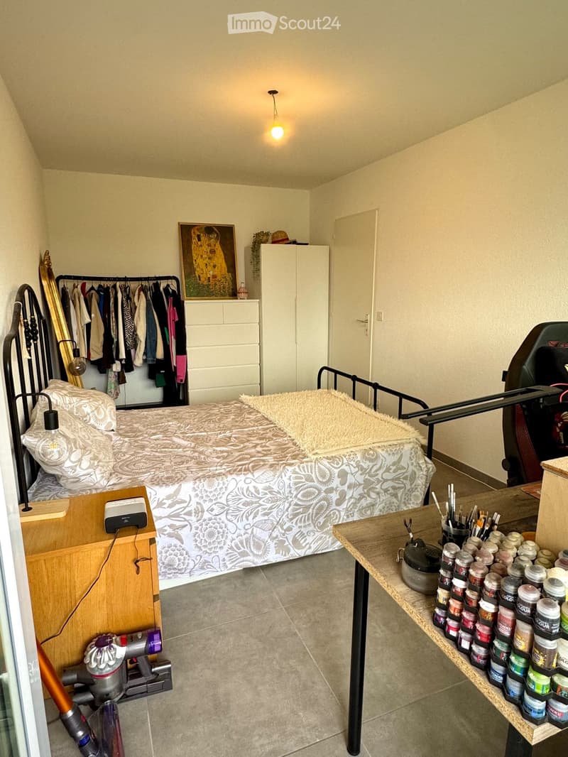 Cozy bedroom with a bed, wardrobe, small table with art supplies, and a framed picture on the wall.