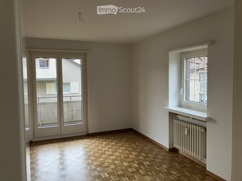Empty room with wooden parquet flooring, white walls, a window with a radiator below, and a glass door leading to a balcony.