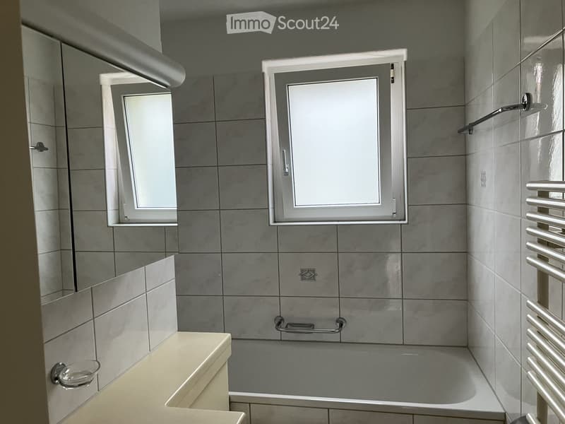 Modern bathroom with grey tiled walls, a bathtub, a large mirror, a radiator, and a small cup on the edge of the sink.