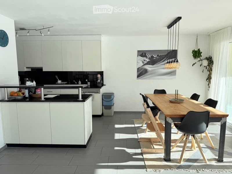 Modern kitchen with white cabinets, black countertops, a wooden dining table with chairs, pendant lights, and a large mountain-themed picture on the wall.