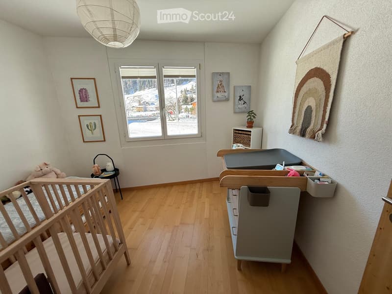 A well-lit nursery with a wooden crib, changing table, storage unit, and various baby items. The room features framed artwork, a colorful rug, and a view of a snowy landscape through large windows.