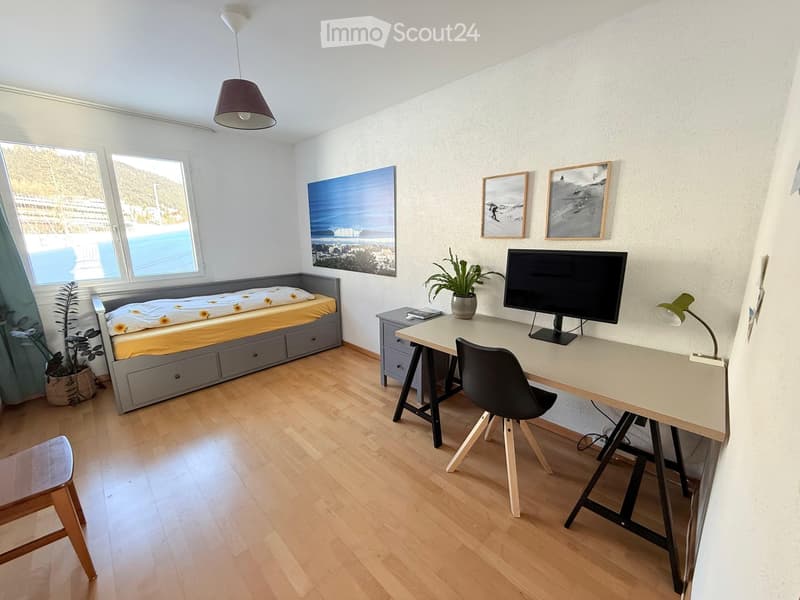 Cozy room with a bed, desk, chair, and a small stool. Features a large window with a view, a hanging lamp, and several framed pictures on the wall.