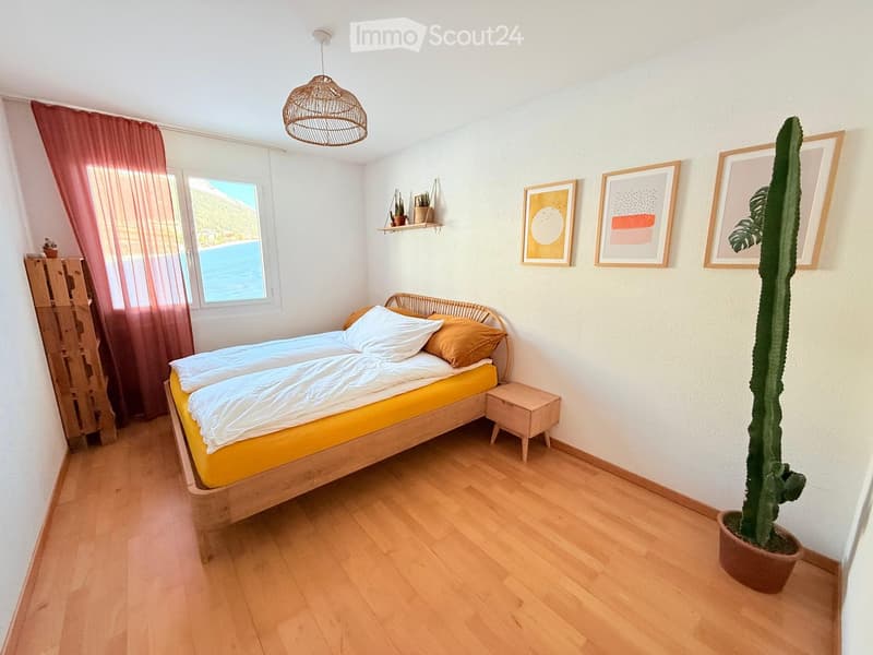 Bright bedroom with wooden floor, large window with view, bed with yellow mattress, nightstand, cactus plant, and wall art.