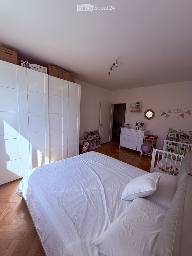 Bright bedroom with a large bed, white wardrobe, dresser, and various toys and shelves.
