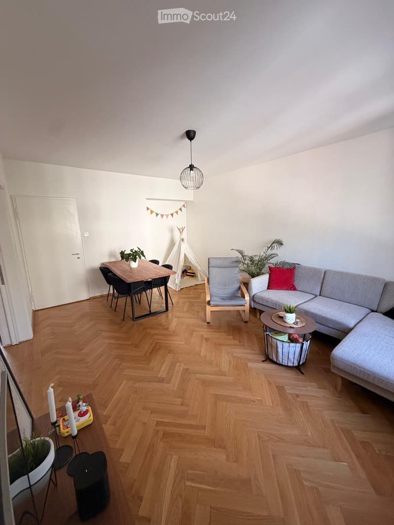 A modern living room with a grey sectional sofa, coffee table, dining table with chairs, pendant light, and decorative plants.