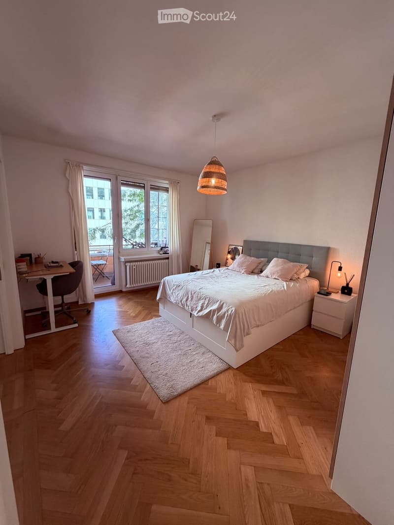 Modern bedroom with wooden flooring, large bed with white bedding, nightstand, desk, and a view through large windows.