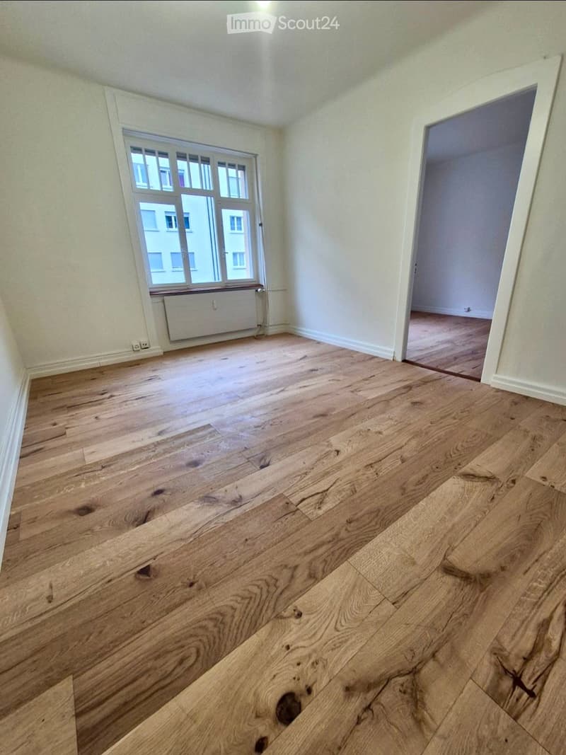 Empty room with wooden flooring, white walls, and a large window with a radiator below it. An open doorway leads to another room with wooden flooring.