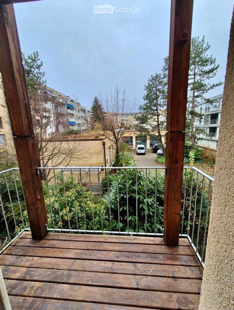Wooden balcony with metal railing, overlooking a residential area with trees and parked cars.