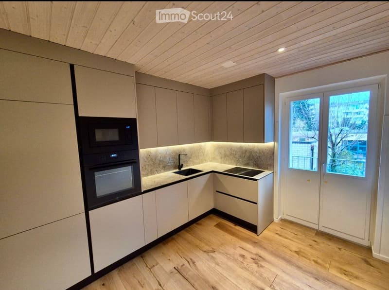 Modern kitchen with light wood flooring, grey cabinets, built-in microwave and oven, sink with faucet, and a window with a view of the outside.