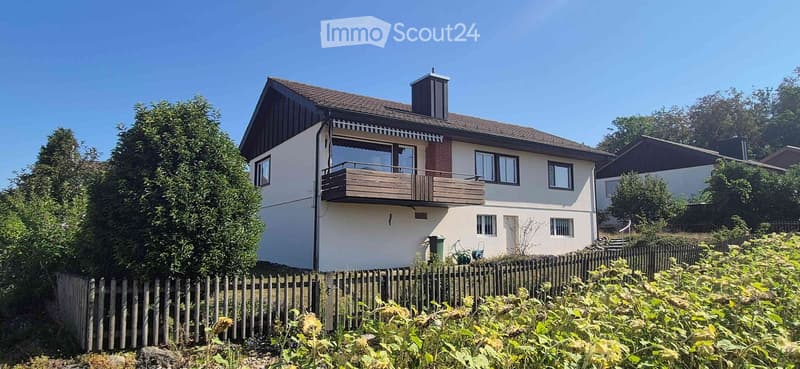 Two-story house with a balcony, surrounded by greenery and a wooden fence.