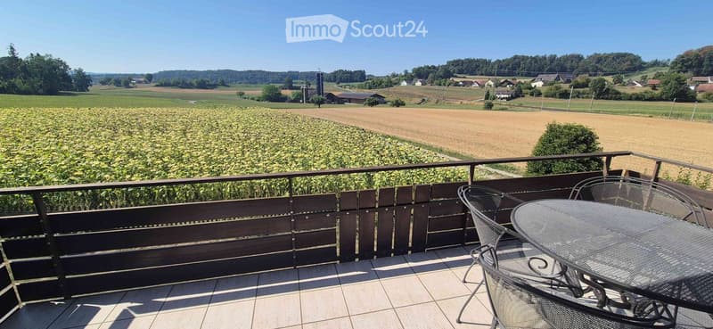 Balcony with a wooden railing, tiled floor, and a view of a rural landscape with fields and houses.