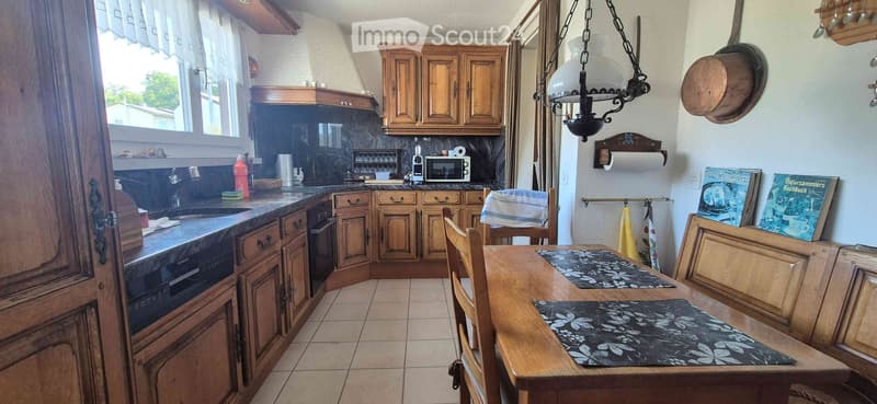 A cozy kitchen with wooden cabinets, a sink, microwave, and a dining table with chairs. The space features a tiled floor and a hanging chandelier.