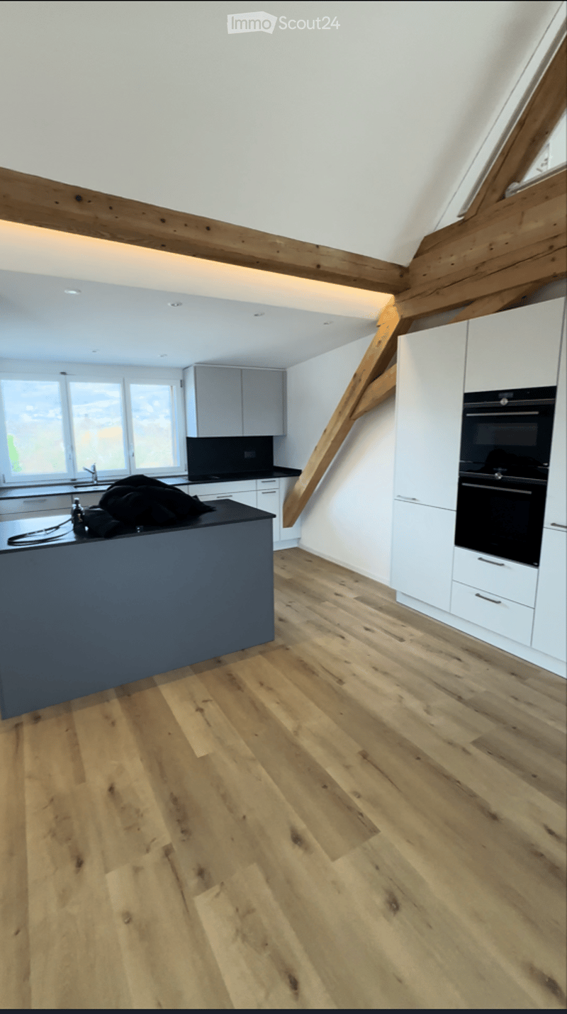 Modern kitchen with light wooden flooring, white cabinets, a black kitchen island, and a built-in oven. Large windows provide natural light.