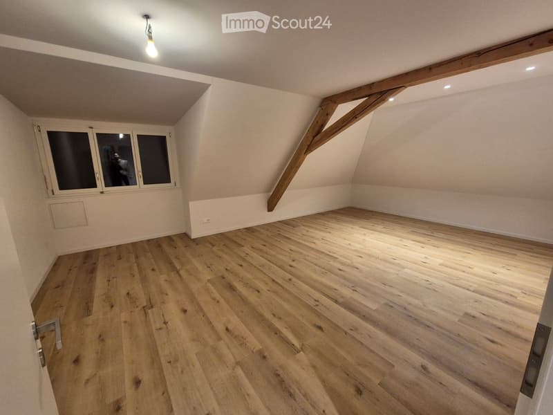 Empty attic room with wooden flooring, exposed wooden beams, and large windows.