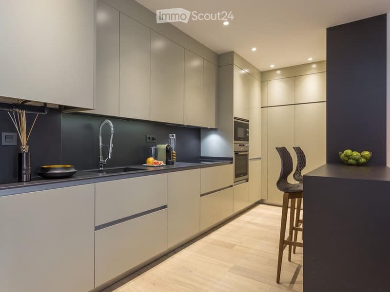 Modern kitchen with light-colored cabinets, dark countertops, and a central island with bar stools. Features include a sink, faucet, oven, and decorative items on the counters.