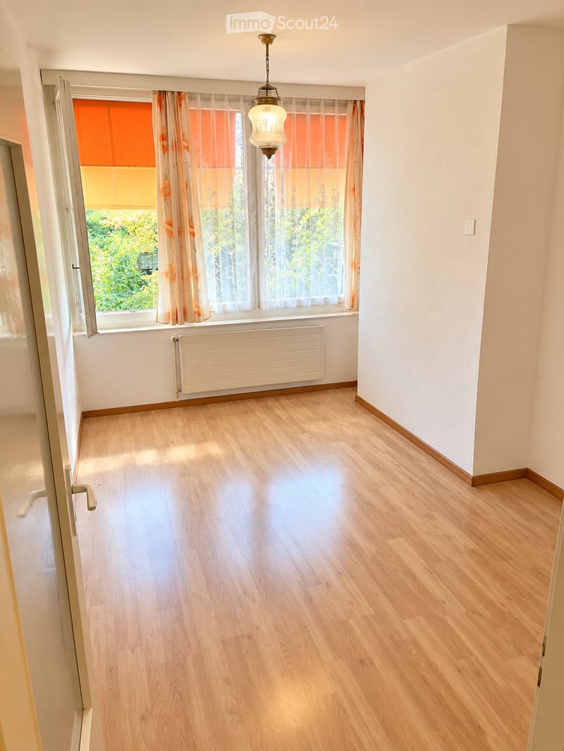 Empty room with light wooden flooring, white walls, a radiator, and a window with curtains. The room is well-lit with natural light coming through the window.