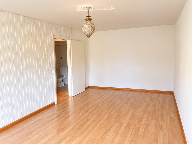 Empty room with light wooden flooring, white walls, and a decorative hanging light fixture. An open doorway leads to a small bathroom with a toilet visible.