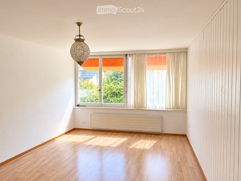 Empty room with light wooden flooring, white walls, large windows with white curtains, and a decorative hanging light fixture.