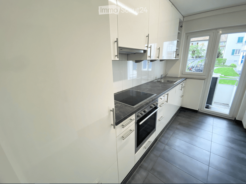 Modern kitchen with white cabinets, dark countertops, induction cooktop, oven, and sink. Large window offers a view of the garden.