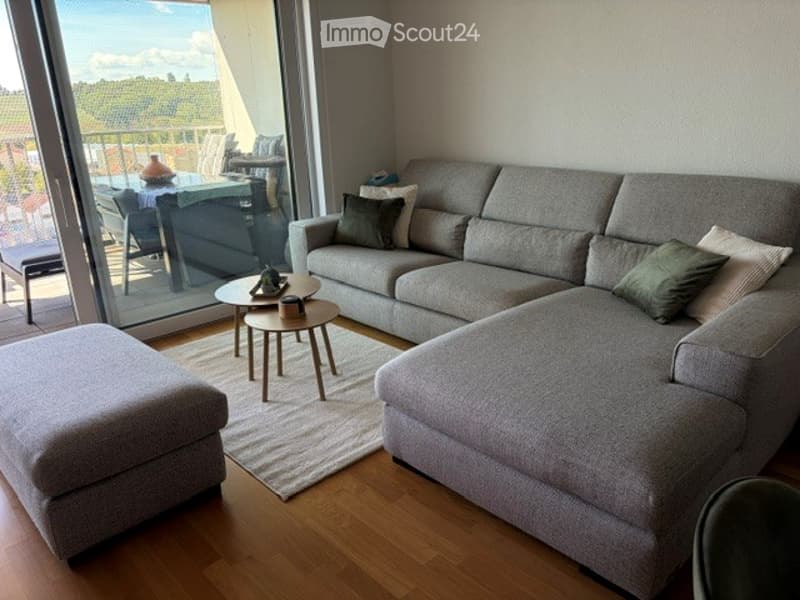 Modern living room with grey sectional sofa, ottoman, coffee table, side table, and a view through large windows.