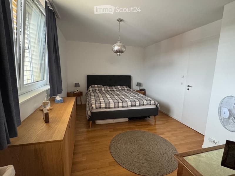 Cozy bedroom with wooden flooring, a large bed with a plaid blanket, a wooden dresser, a round woven rug, and a fan near the door.
