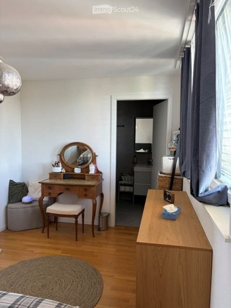 A small, cozy room with a wooden floor, a vanity with a mirror, a small table, and a few stools. The room has a window with curtains and a small seating area with a cushioned stool.