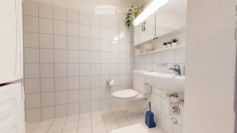 A small, clean bathroom with white tiled walls and floor, featuring a toilet, sink with a modern faucet, a mirror above the sink, shelves with decorative items, and a blue mop on the floor.