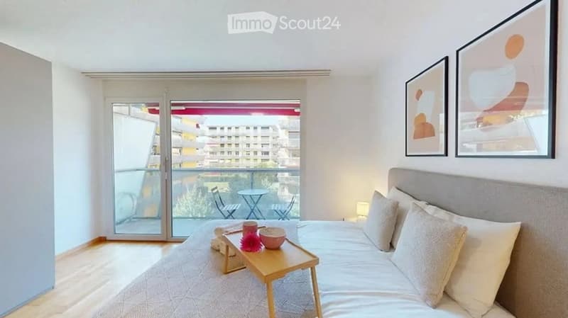 Modern bedroom with a large bed, nightstand, small table with decorative items, and access to a balcony with outdoor furniture.