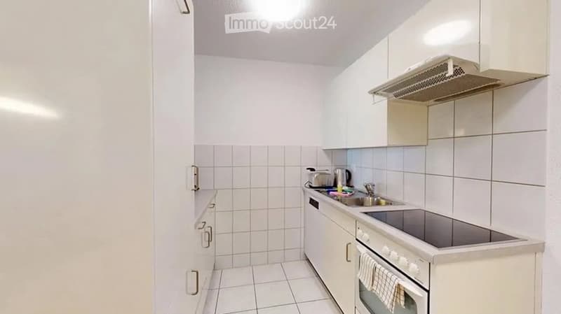 Modern kitchen with white tiled walls and floor, white cabinets, built-in oven, sink, and range hood.