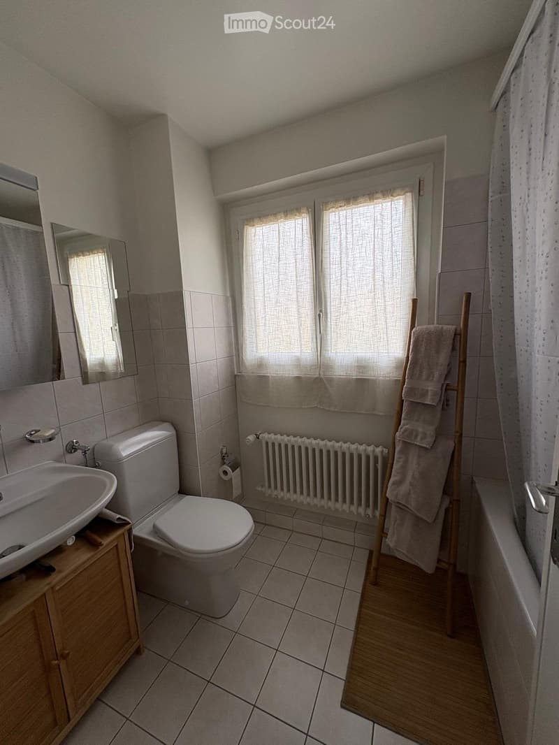 Bathroom with a toilet, sink, mirror, radiator, towel rack with towels, and a bathtub. The room has tiled walls and floor, with a window covered by a curtain.
