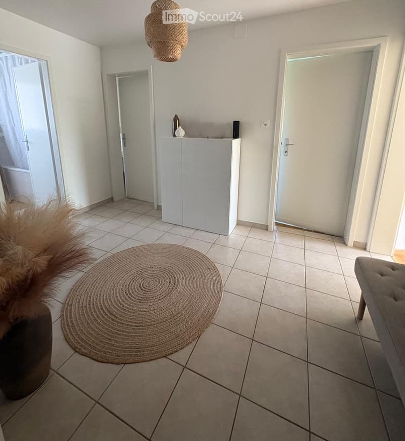 A modern, minimalist hallway with tiled flooring, a round woven rug, a small white console table with decorative items, and a pendant light fixture.