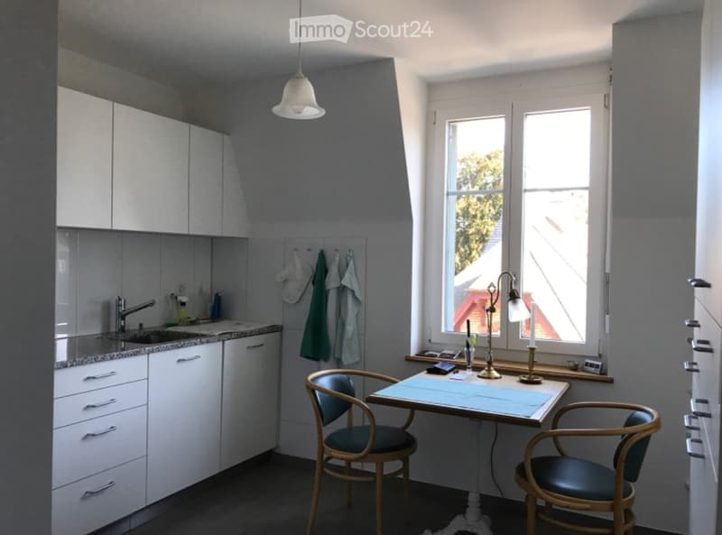 A small, modern kitchen with white cabinets, a sink, a small table with two chairs, and a window with a view of the outside.