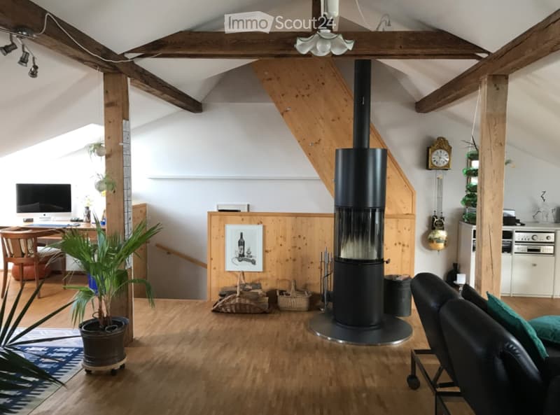 A spacious living room with wooden beams, a central black fireplace, a sofa, a chair, a small table, a screen, and a potted plant.