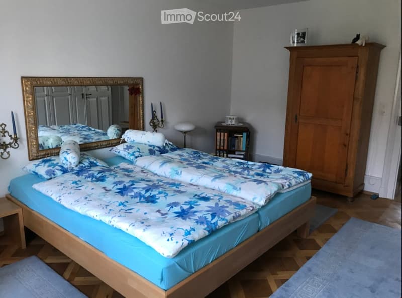 A well-furnished bedroom with a wooden bed, blue floral bedding, a large mirror with a gold frame, a wooden wardrobe, a small table with books, and a stool.