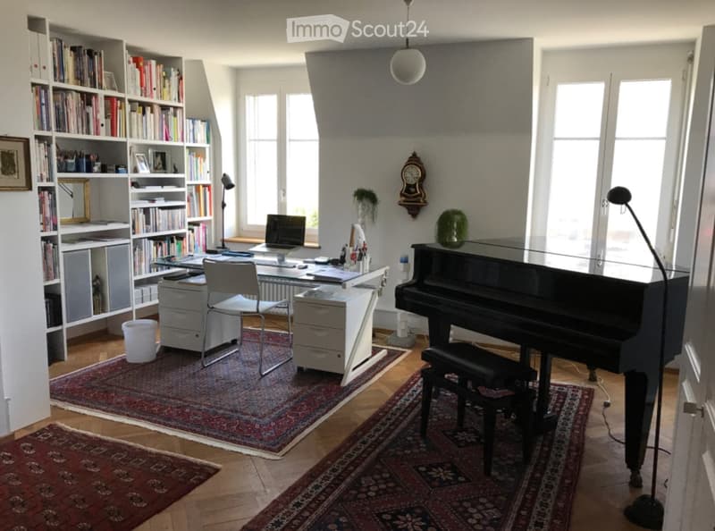Brightly lit room with wooden flooring, large windows, bookshelf filled with books, desk with laptop, chair, piano, and decorative items.