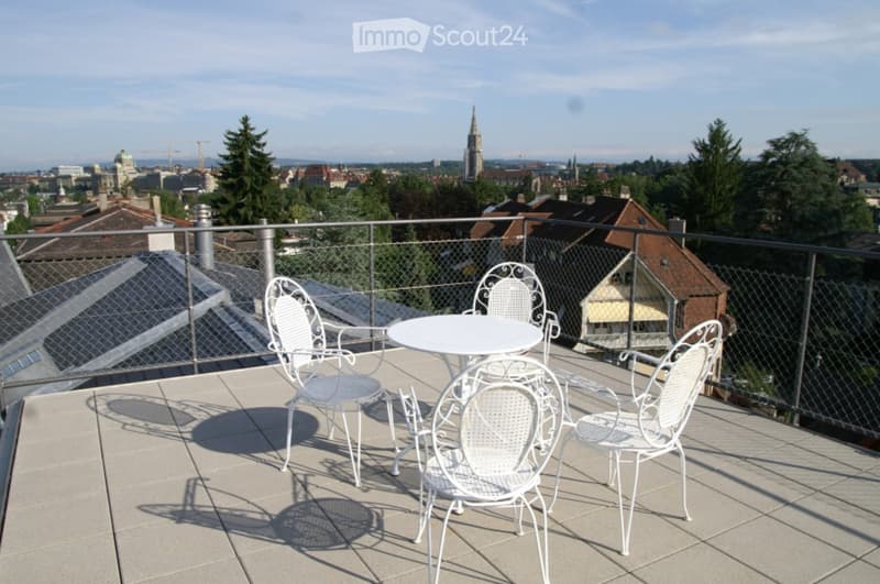 A rooftop terrace with a white metal table and four matching chairs, overlooking a cityscape with visible buildings and trees.
