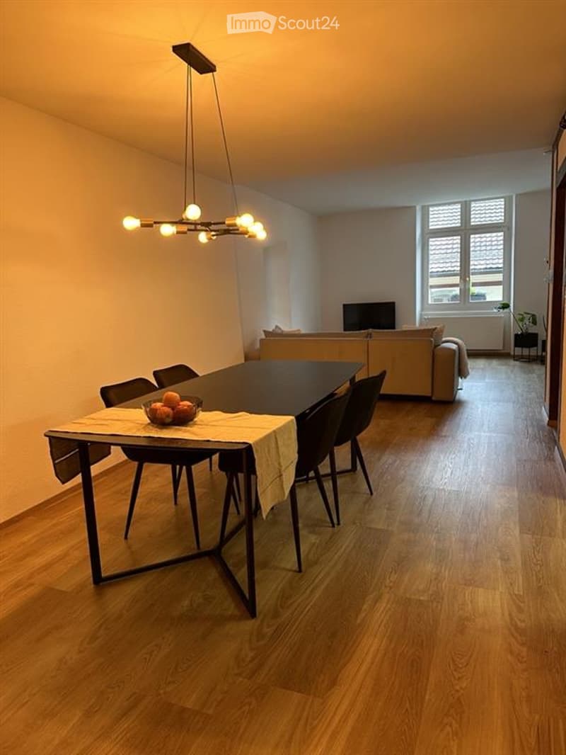 Open-plan living and dining area with wooden flooring, a modern chandelier, a dining table with chairs, a sofa, and a television in the background.