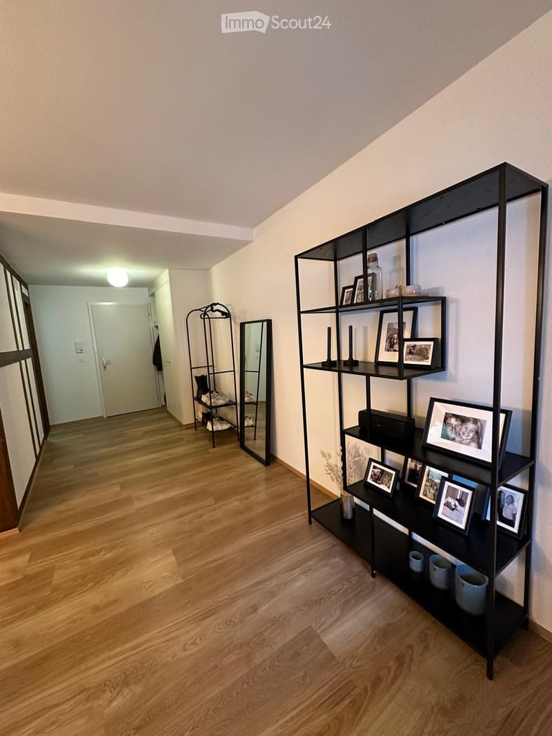 A well-lit hallway with wooden flooring, a black shelving unit with framed photos and decorative items, a mirror, and a coat rack with shoes and jackets.