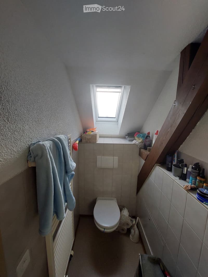 Small bathroom with a toilet, tiled walls, a window, a towel hanging on the left, and cleaning supplies on the right shelf.