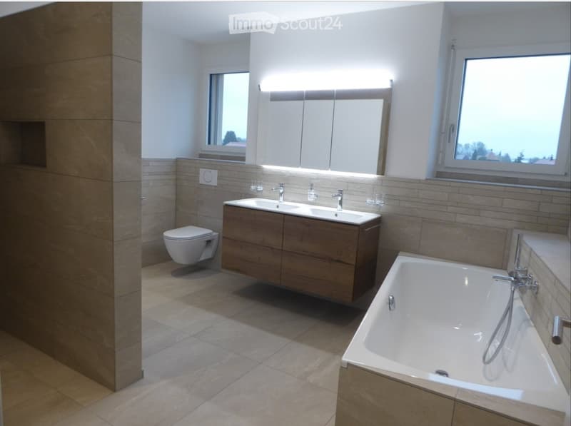 Modern bathroom with a bathtub, toilet, double sink, and large mirror. Light-colored tiles and wooden vanity.