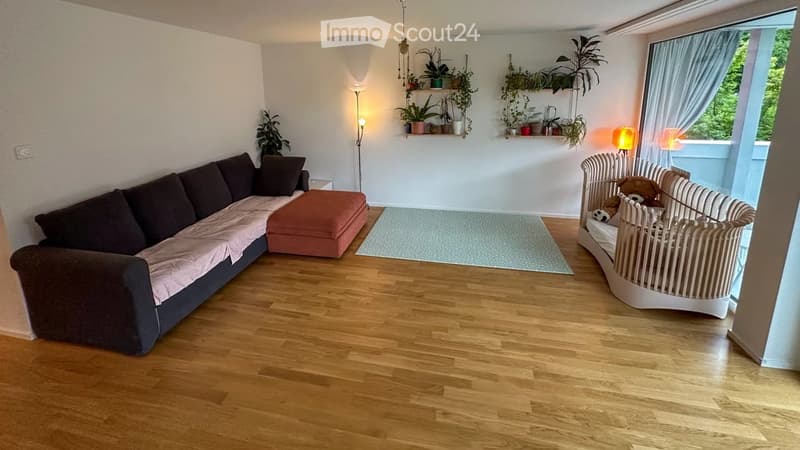 Living room with wooden flooring, a dark brown sectional sofa with pink throw, an orange ottoman, a white baby crib with stuffed animals, floor lamp, table lamp, and wall-mounted shelves with plants.