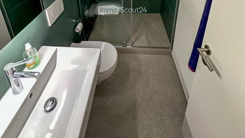 Modern bathroom with a white sink, chrome faucet, toilet, and a glass-enclosed shower area. Green tiles on one wall and beige tiles on the floor.