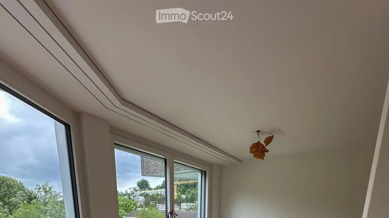 Empty room with large windows offering a view of the city and greenery, featuring a modern ceiling design and a decorative leaf-shaped light fixture.