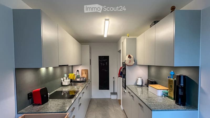 Modern kitchen with white cabinets, granite countertops, and various appliances including a coffee maker, toaster, and cutting board. A sink with a faucet is visible, along with a light fixture above the counter.