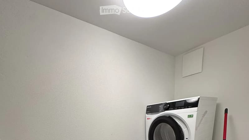 A small, clean room with a modern washing machine, white walls, and a skylight window on the ceiling.