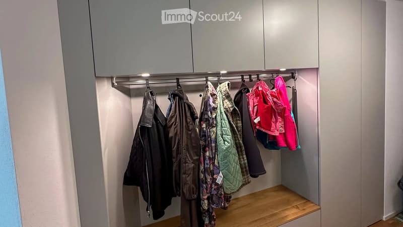 Modern hallway with built-in storage cabinets, a coat rack holding several jackets, and a small wooden bench at the bottom.