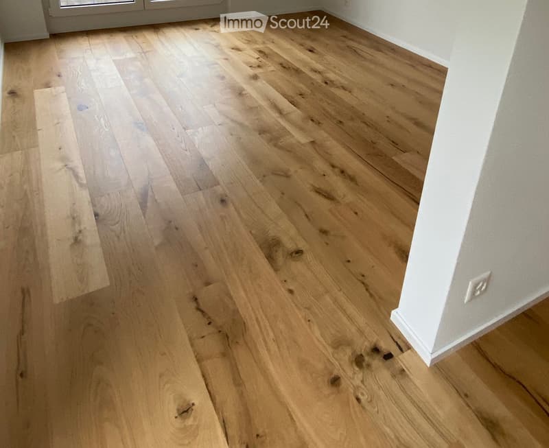 Empty room with light wooden flooring and white walls, showing natural wood grain and subtle imperfections.